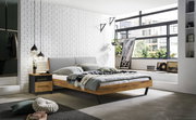 Modernes Bettgestell aus Holz mit grauem Kopfteil, seitliche Perspektive, in einem stilvoll eingerichteten Schlafzimmer mit großen Fenstern und dekorativen Elementen.