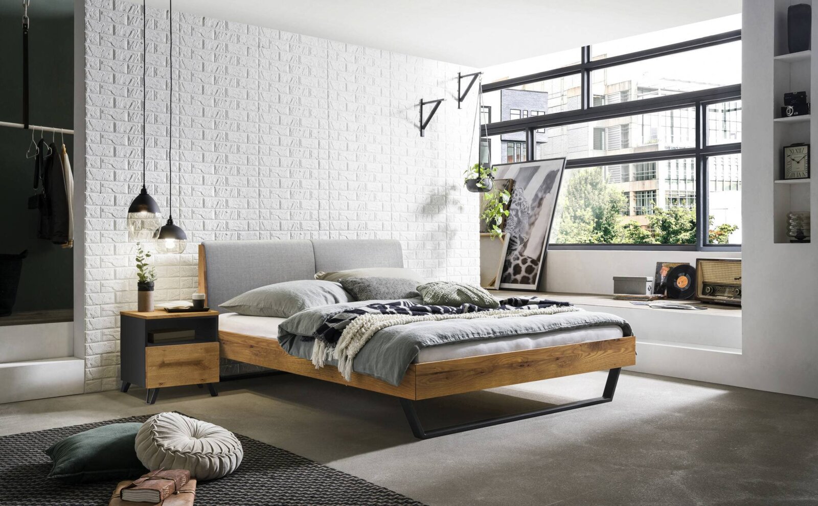 Modernes Bettgestell aus Holz mit grauem Kopfteil, seitliche Perspektive, in einem stilvoll eingerichteten Schlafzimmer mit großen Fenstern und dekorativen Elementen.