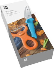 Koch-Set WILLY MIA FRED, bestehend aus einem blauen Messer und einem orangefarbenen Schäler mit Affenmotiv, auf einer Holzoberfläche mit geschnittenen Karotten, aus der Vogelperspektive.