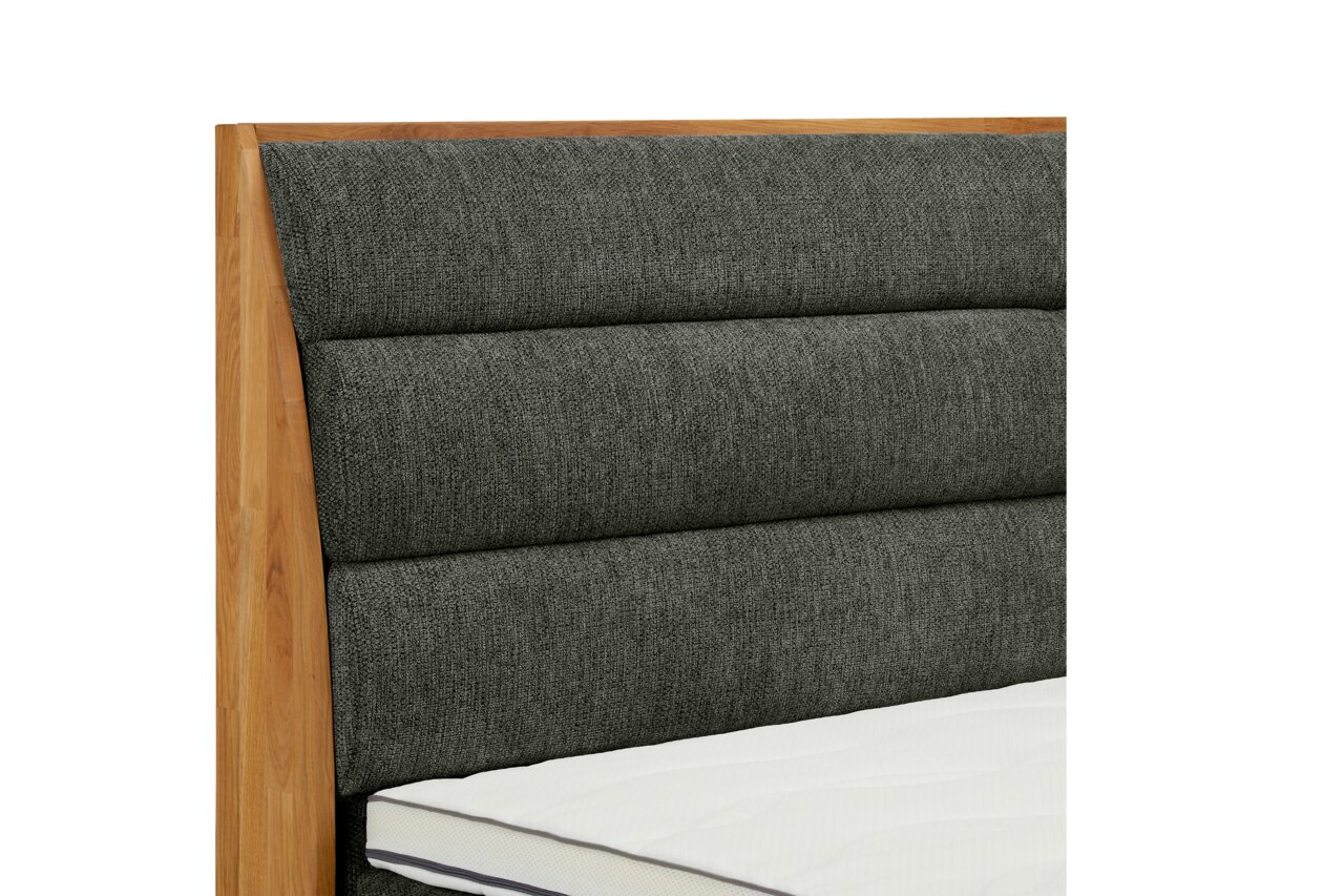 Nahaufnahme eines Boxspringbettes mit grauem, gepolstertem Kopfteil und Holzrahmen, seitliche Perspektive