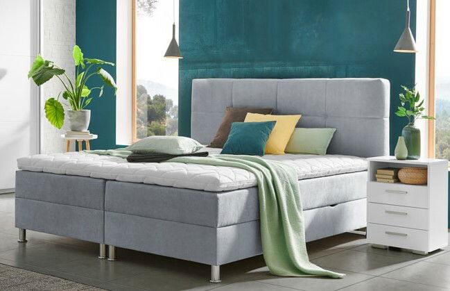 Modernes Boxbett in einem stilvollen Schlafzimmer, frontal fotografiert, mit grauem Polsterkopfteil, dekorativen Kissen und einer grünen Decke, umgeben von Pflanzen und Nachttisch.