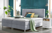 Modernes Boxbett in einem stilvollen Schlafzimmer, frontal fotografiert, mit grauem Polsterkopfteil, dekorativen Kissen und einer grünen Decke, umgeben von Pflanzen und Nachttisch.