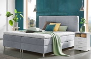 Modernes Boxbett in einem stilvollen Schlafzimmer, frontal fotografiert, mit grauem Polsterkopfteil, dekorativen Kissen und einer grünen Decke, umgeben von Pflanzen und Nachttisch.