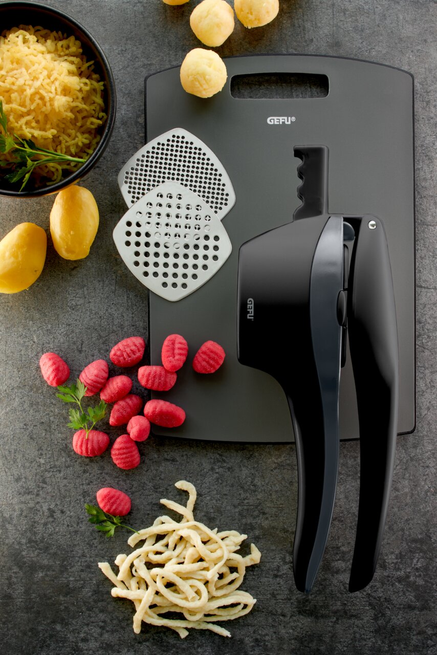 Kartoffel- und Spätzlepresse mit zwei Einsätzen, umgeben von Kartoffeln, Spätzle und Gnocchi, aus der Vogelperspektive.