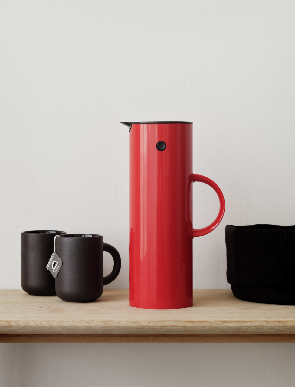 Stelton Isolierkanne 1l EM77 Rote Isolierkanne mit einem Liter Fassungsvermögen, seitlich auf einem Regal neben zwei schwarzen Tassen und einem schwarzen Behälter platziert.