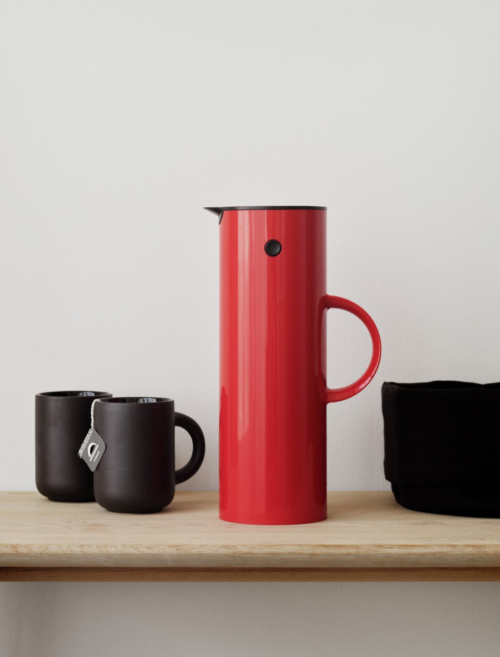 Stelton Isolierkanne 1l EM77 Rote Isolierkanne mit einem Liter Fassungsvermögen, seitlich auf einem Regal neben zwei schwarzen Tassen und einem schwarzen Behälter platziert.