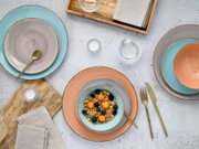 Draufsicht auf ein farbiges Keramikgeschirr-Set mit Tellern und Schalen in Türkis, Grau und Terrakotta auf heller Tischplatte, mit goldfarbenem Besteck, Gläsern, Holztablett und Leinenservietten; eine Schale ist mit Müsli und Beeren gefüllt.
