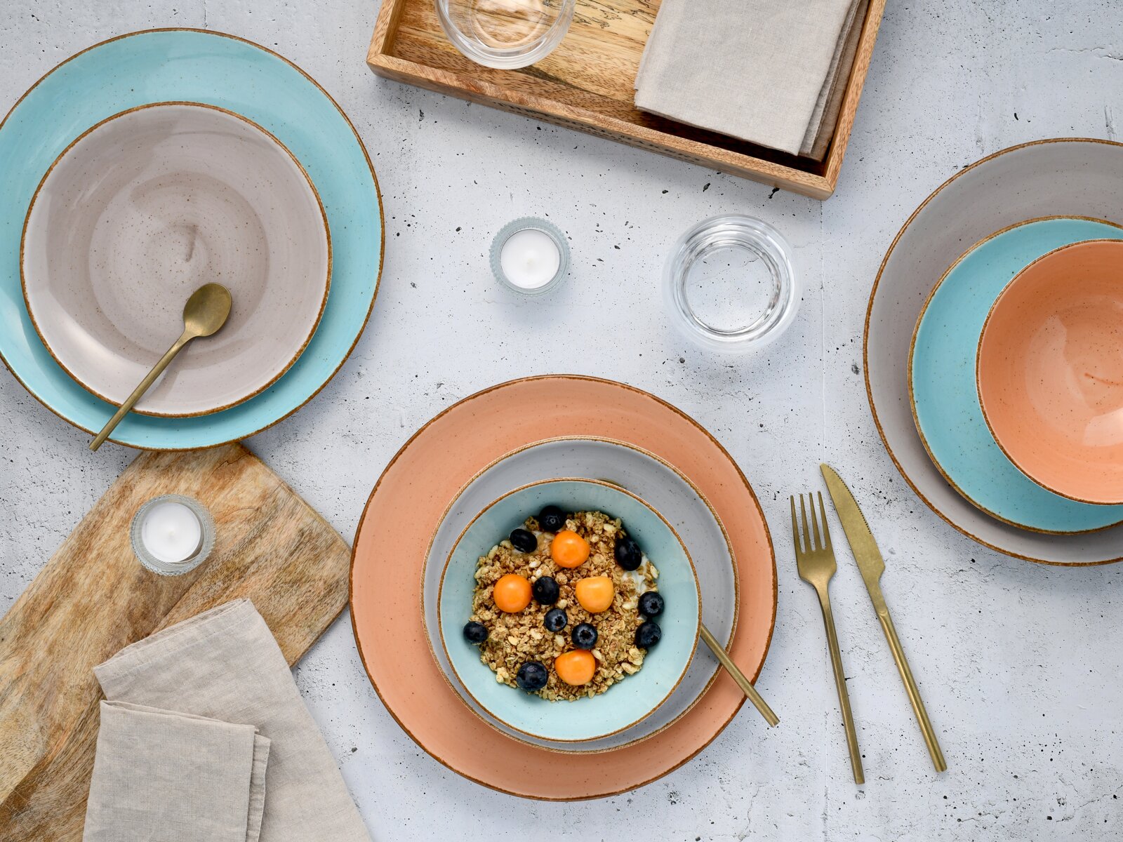 Draufsicht auf ein farbiges Keramikgeschirr-Set mit Tellern und Schalen in Türkis, Grau und Terrakotta auf heller Tischplatte, mit goldfarbenem Besteck, Gläsern, Holztablett und Leinenservietten; eine Schale ist mit Müsli und Beeren gefüllt.