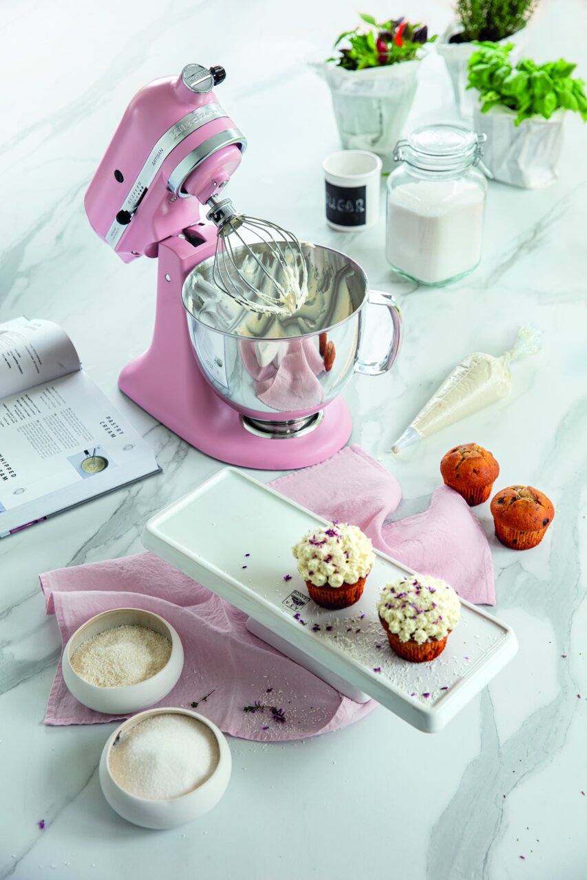Rosa Küchenmaschine 4.8L Artisan mit Rührbesen, umgeben von Backzutaten und Cupcakes, aus der Vogelperspektive.