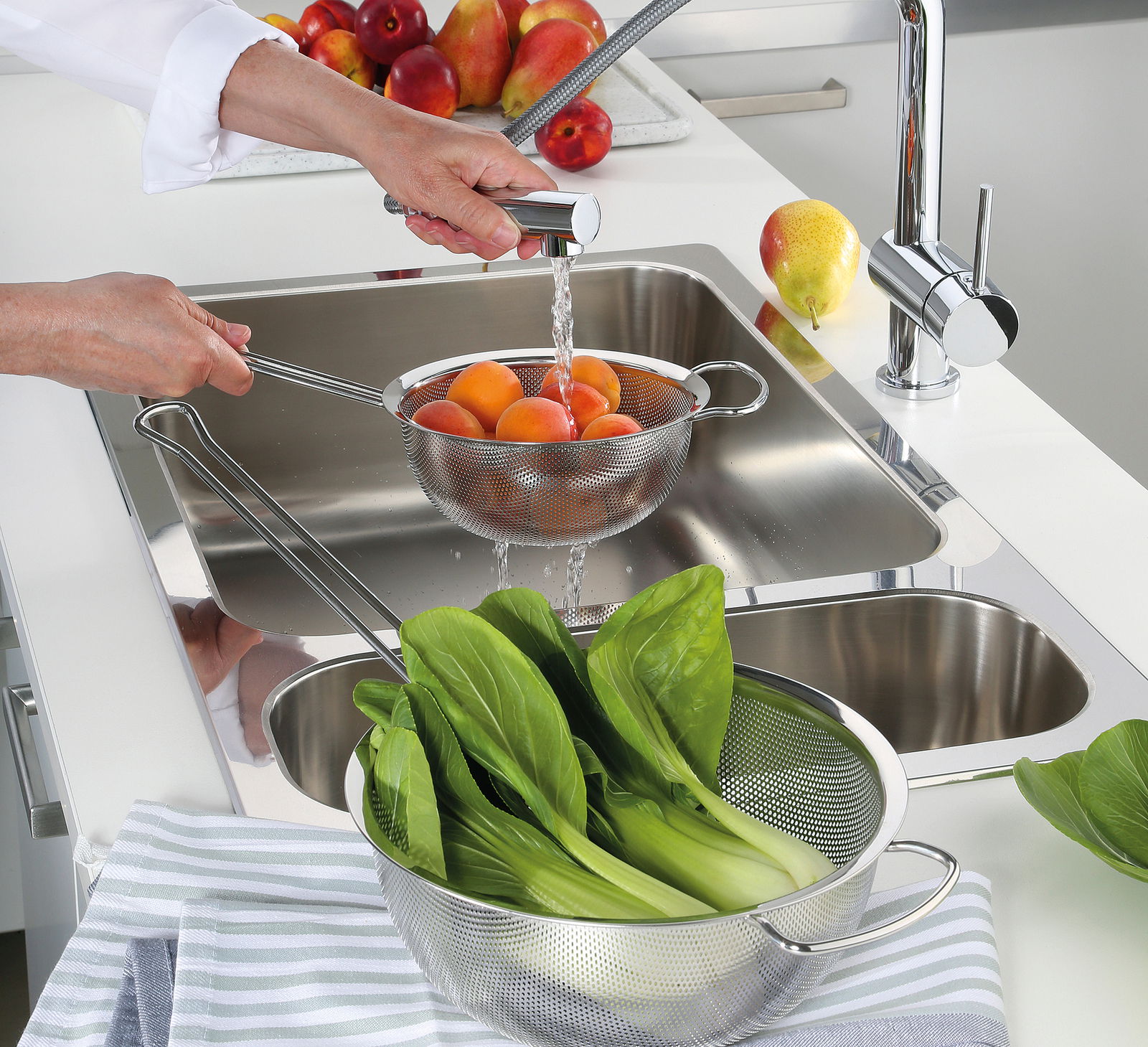 Küchensieb DELUXE, 22 cm, in einer modernen Küche, gefüllt mit frischen Aprikosen, während Wasser darüber fließt. Perspektive von oben auf das Sieb und die Spüle.