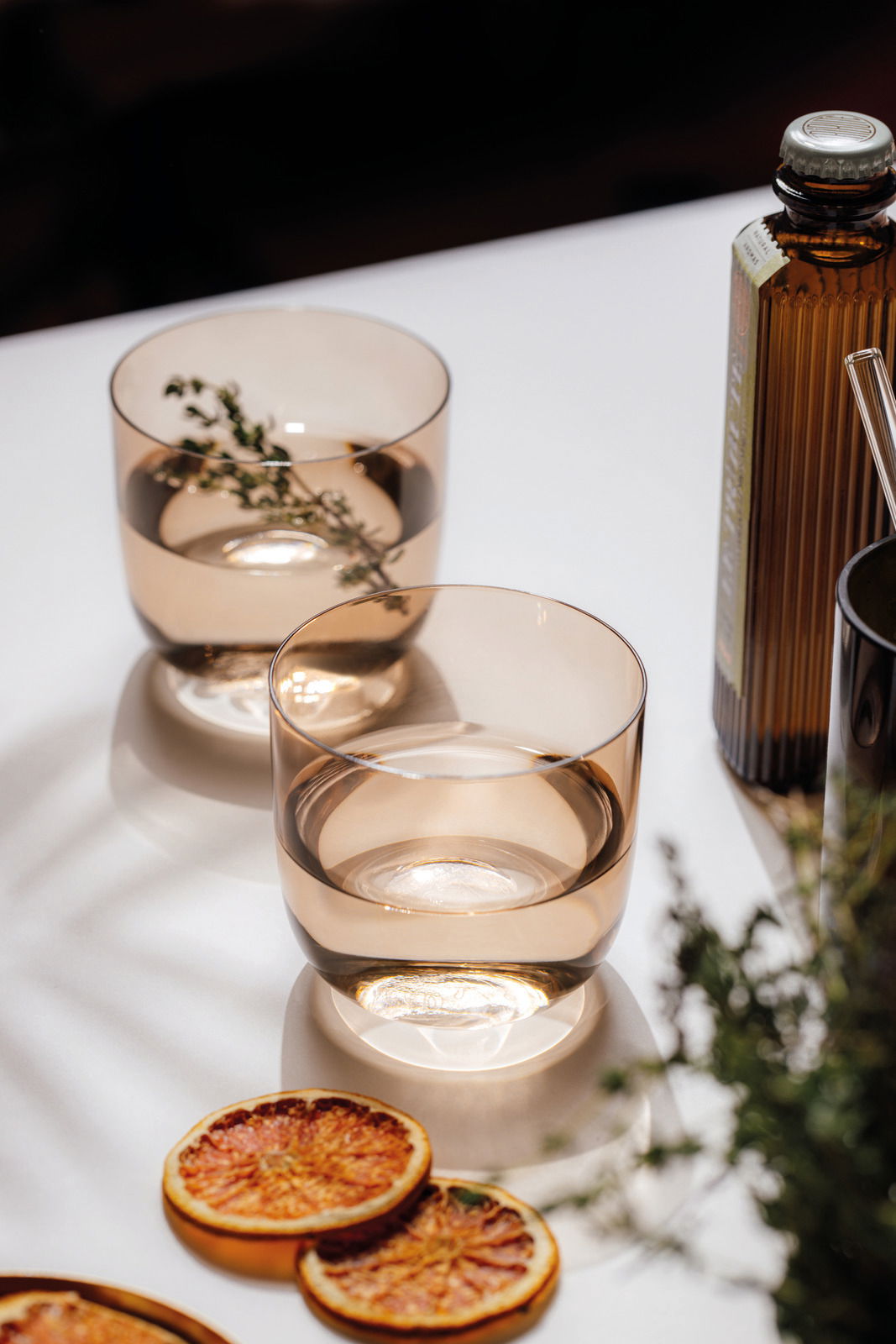 Zwei elegante Wassergläser in warmem Licht, gefüllt mit Wasser und einem Zweig Thymian, auf einem Tisch mit getrockneten Orangenscheiben und einer Flasche im Hintergrund. Perspektive von oben.