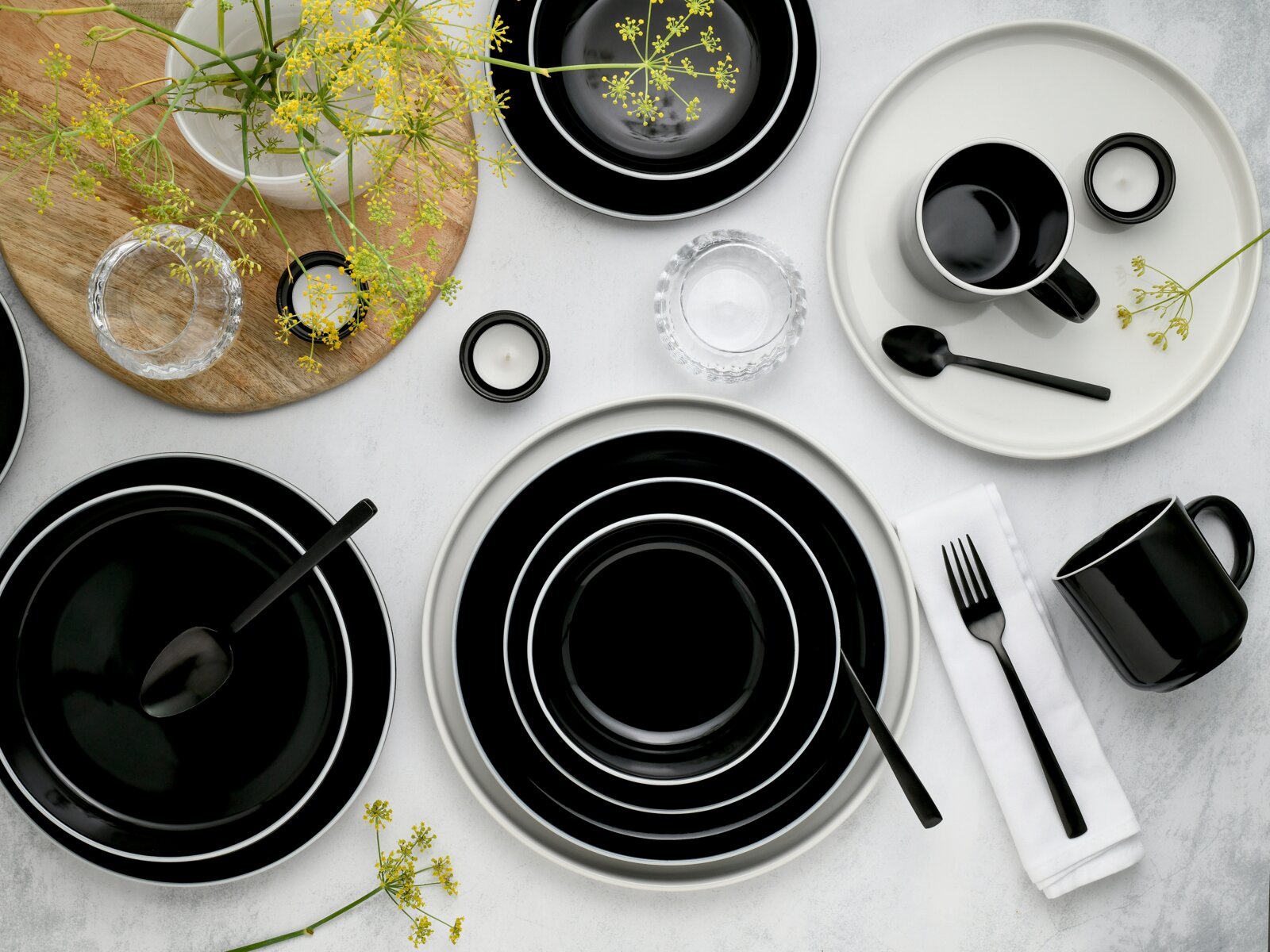 Draufsicht auf ein modernes schwarz-weißes Geschirrset mit Tellern und Schalen, schwarzen Tassen und Besteck, dekoriert mit gelben Blumen auf hellem Untergrund.