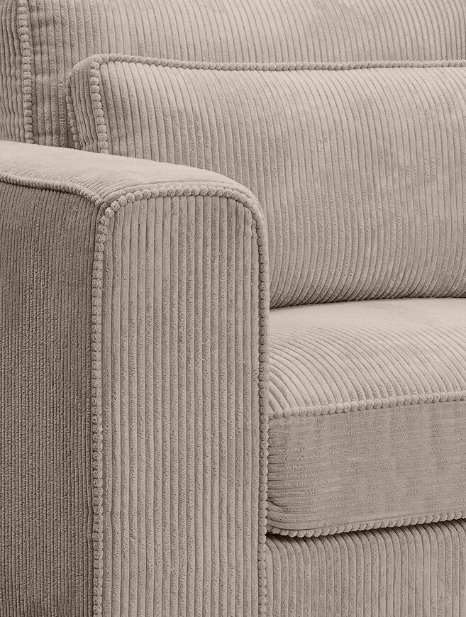 Nahaufnahme eines beigen Cord-Sofas mit geripptem Bezug, breiter Armlehne und Kissen mit dekorativer Paspelierung, fotografiert aus schräger Seitenansicht von vorne.