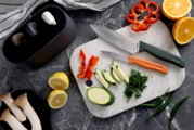 Bovenaanzicht van een witte marmeren snijplank met twee keukenmessen met groene en oranje handgreep, gesneden paprika, courgette en peterselie, met citrusvruchten en chilipepers op een donker werkblad.