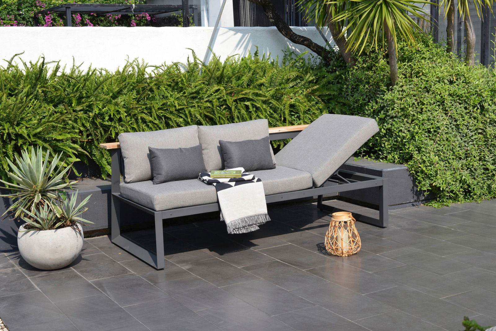 Moderne graue Sitzbank mit verstellbarer Rückenlehne, dekoriert mit Kissen und Decke, auf einer Terrasse mit Pflanzen im Hintergrund, seitliche Perspektive