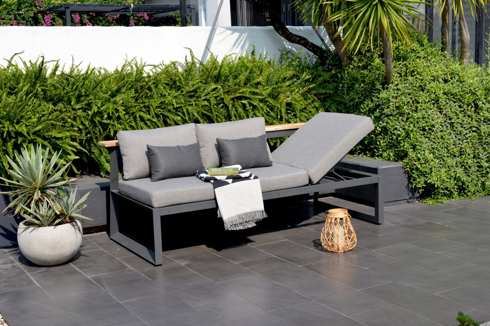 Moderne graue Sitzbank mit verstellbarer Rückenlehne, dekoriert mit Kissen und Decke, auf einer Terrasse mit Pflanzen im Hintergrund, seitliche Perspektive