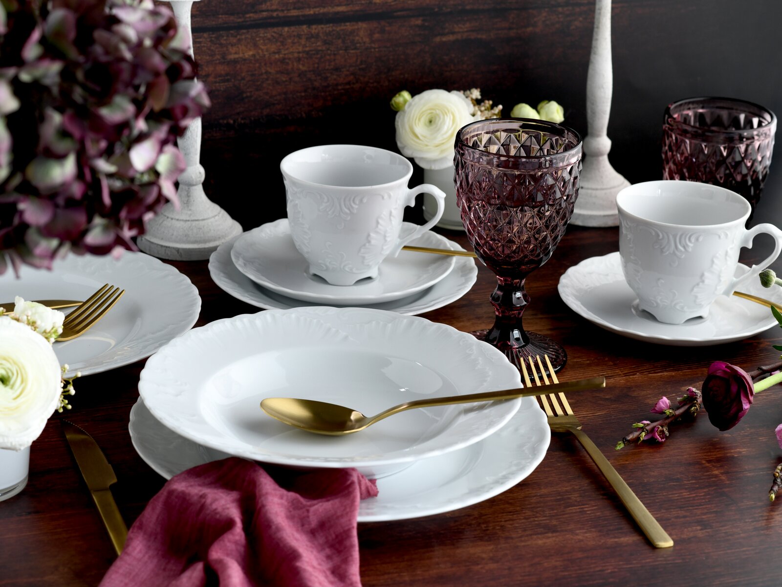 Elegant gedeckter Tisch mit weißem Porzellan-Geschirr (Teller und Tassen mit Relief), goldfarbenem Besteck und violetten Kristallgläsern auf dunkler Holzplatte, fotografiert aus leicht schräger Vorderansicht.