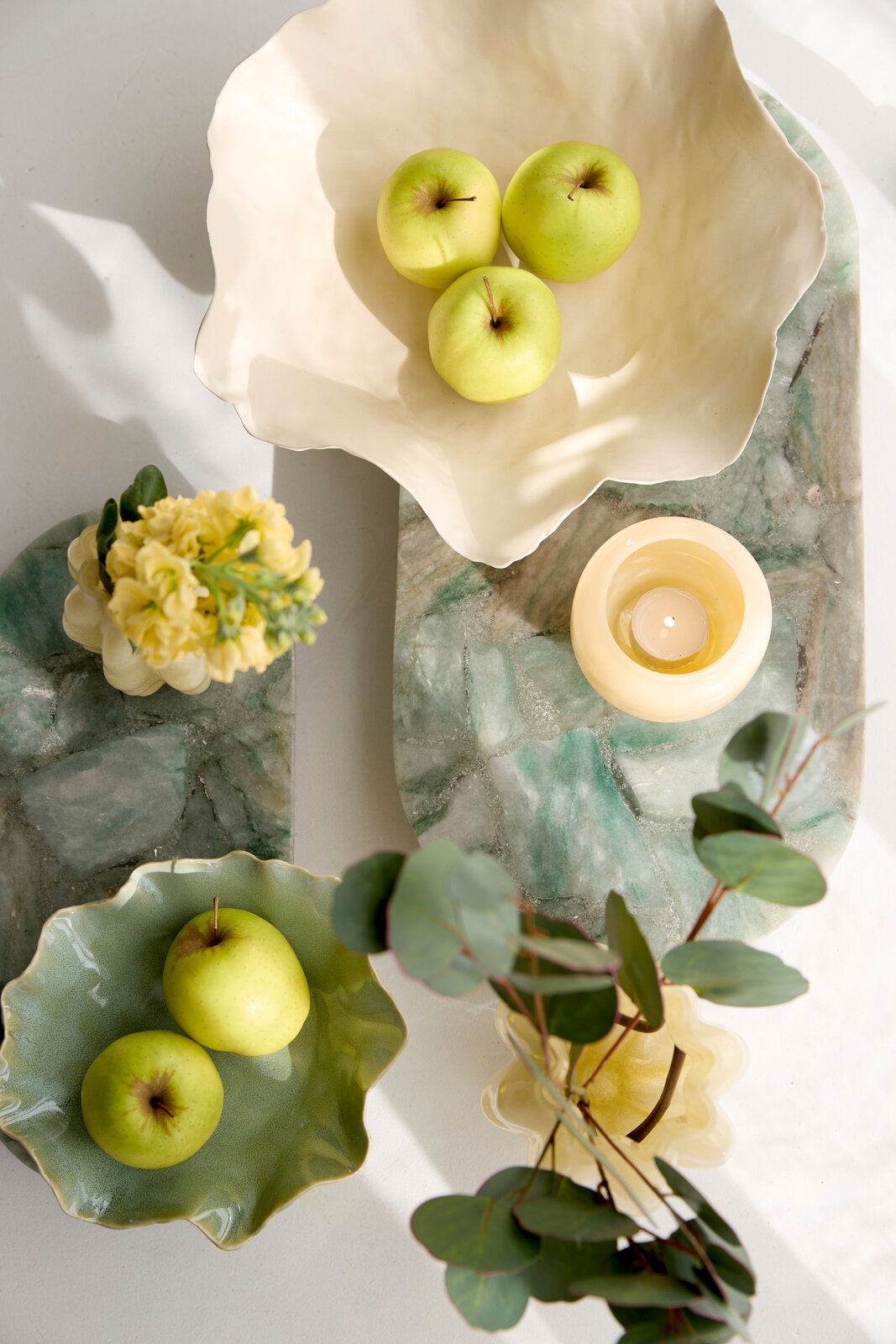 Vogelperspektive auf zwei dekorative Schalen mit grünen Äpfeln, umgeben von Blumen und einer Kerze auf einem grünen Marmorbrett.