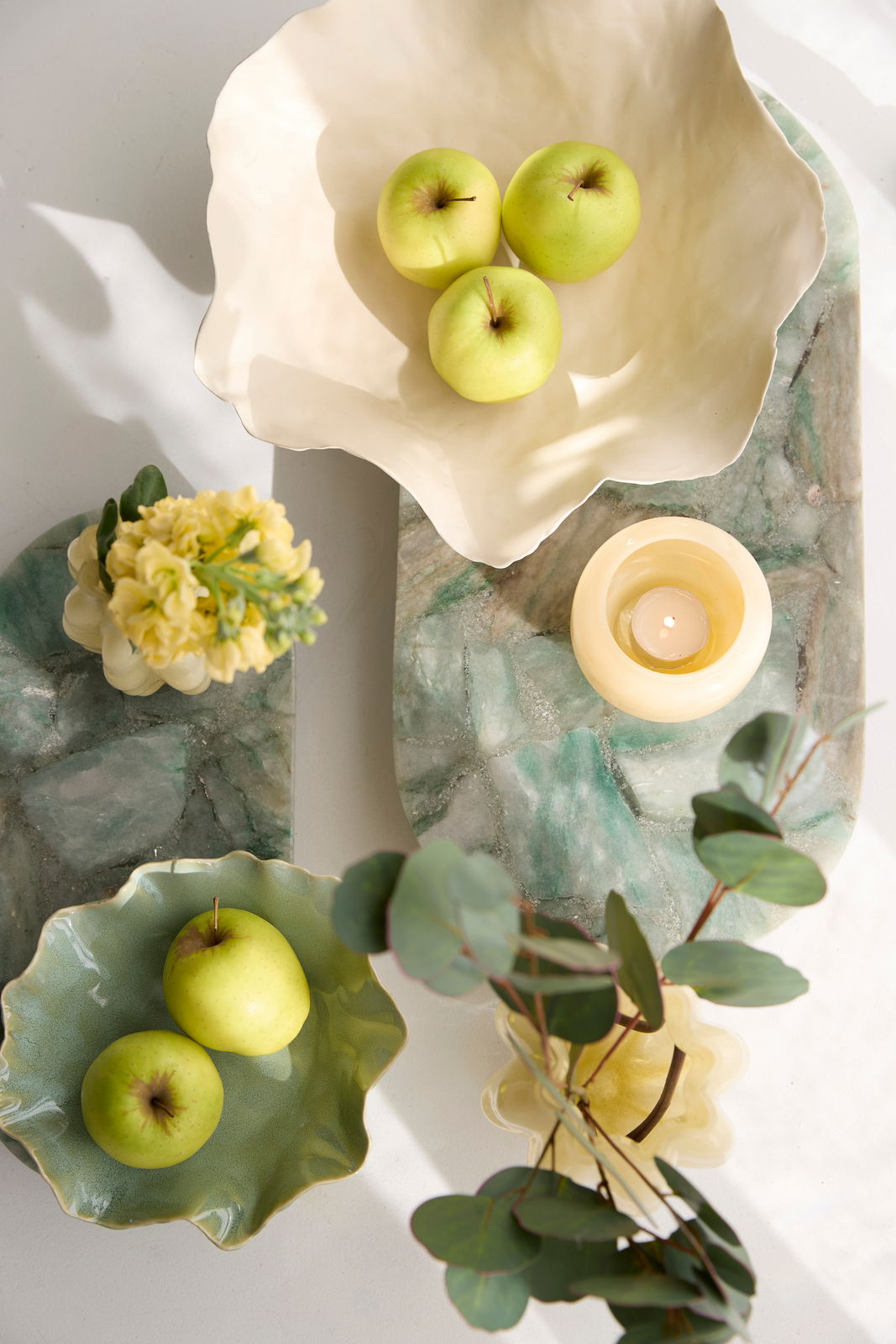 Vogelperspektive auf zwei dekorative Schalen mit grünen Äpfeln, umgeben von Blumen und einer Kerze auf einem grünen Marmorbrett.