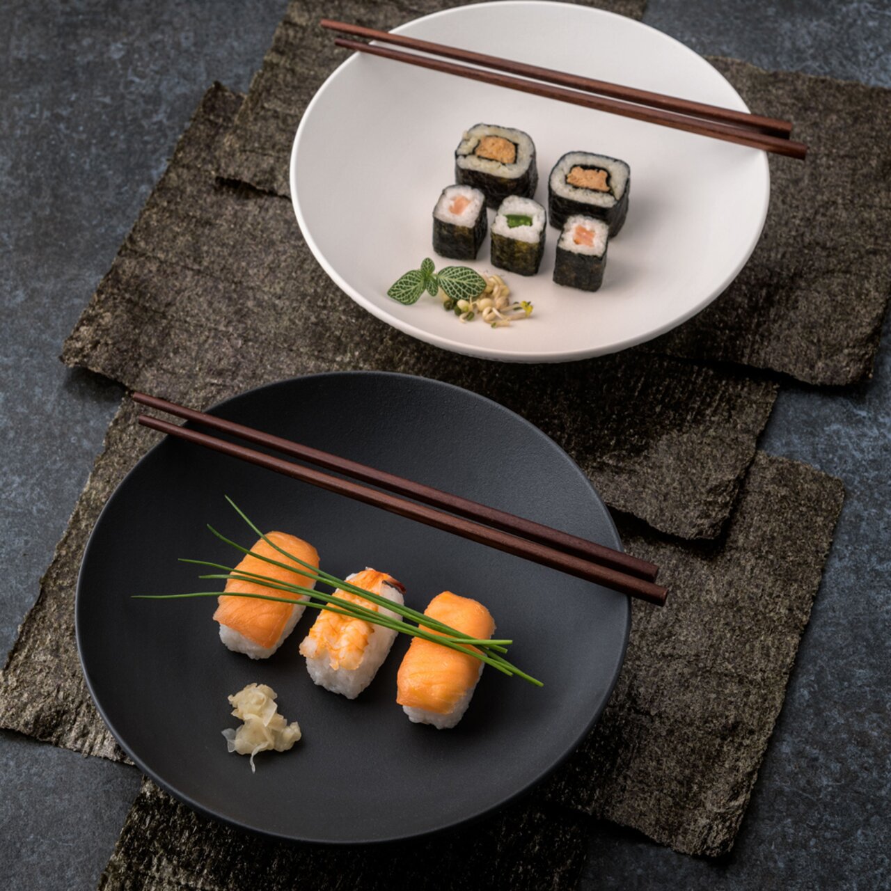 Villeroy & Boch Schale flach MANUFACTURE Zwei Schalen aus der Manufacture-Serie, eine in Schwarz mit Sushi und eine in Weiß mit Maki-Rollen, aus der Vogelperspektive fotografiert, mit Essstäbchen auf dunklem Untergrund.
