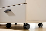 Nahaufnahme eines Rollcontainers mit zwei Schubladen und schwarzen Rollen auf einem Holzboden, seitliche Perspektive.