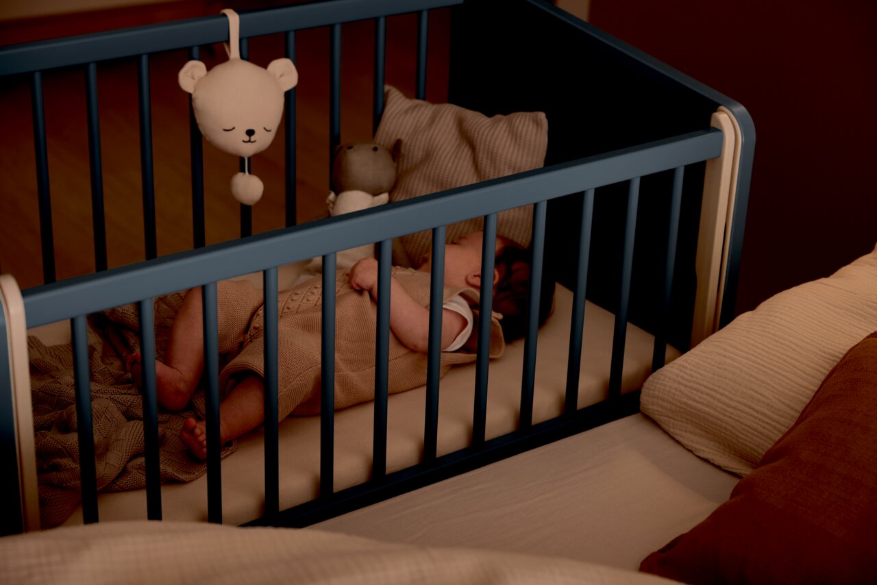 Een baby slaapt in een blauw bijzetbedje dat aan een groter bed is vastgemaakt. Aanzicht van bovenaf.