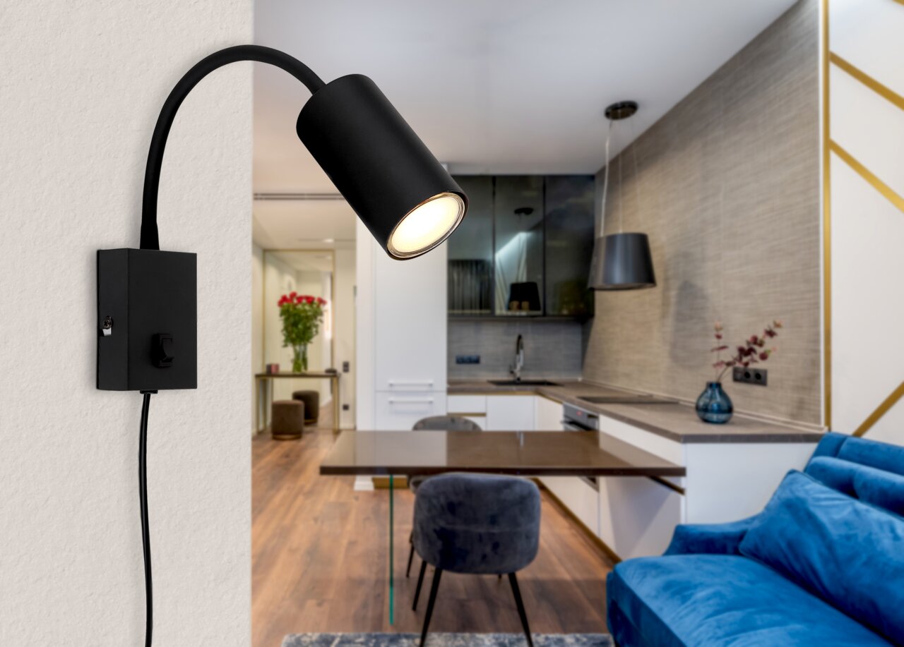 Schwarze, matte Wandleuchte aus Metall, seitliche Perspektive, montiert an einer Wand in einem modernen Wohnzimmer mit offener Küche.