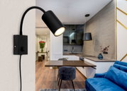 Zwarte, matte wandlamp van metaal, gezien vanaf de zijkant, bevestigd aan een muur in een moderne woonkamer met open keuken.