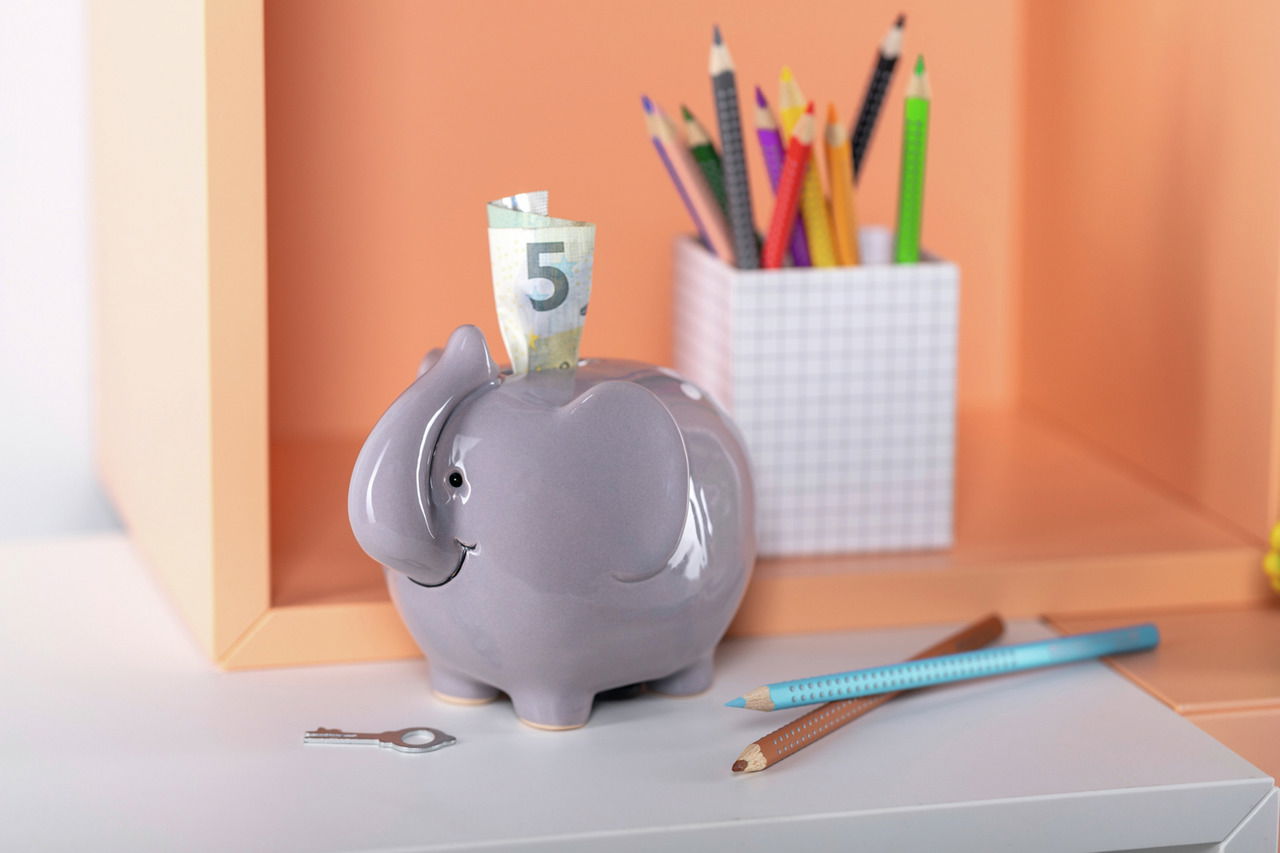 Graue Elefanten-Spardose mit einem Geldschein, seitliche Perspektive, im Hintergrund bunte Stifte.