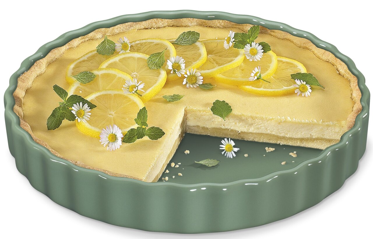 Tortenform mit einem Zitronenkuchen, dekoriert mit Zitronenscheiben und Blüten, aus einer leicht seitlichen Perspektive.