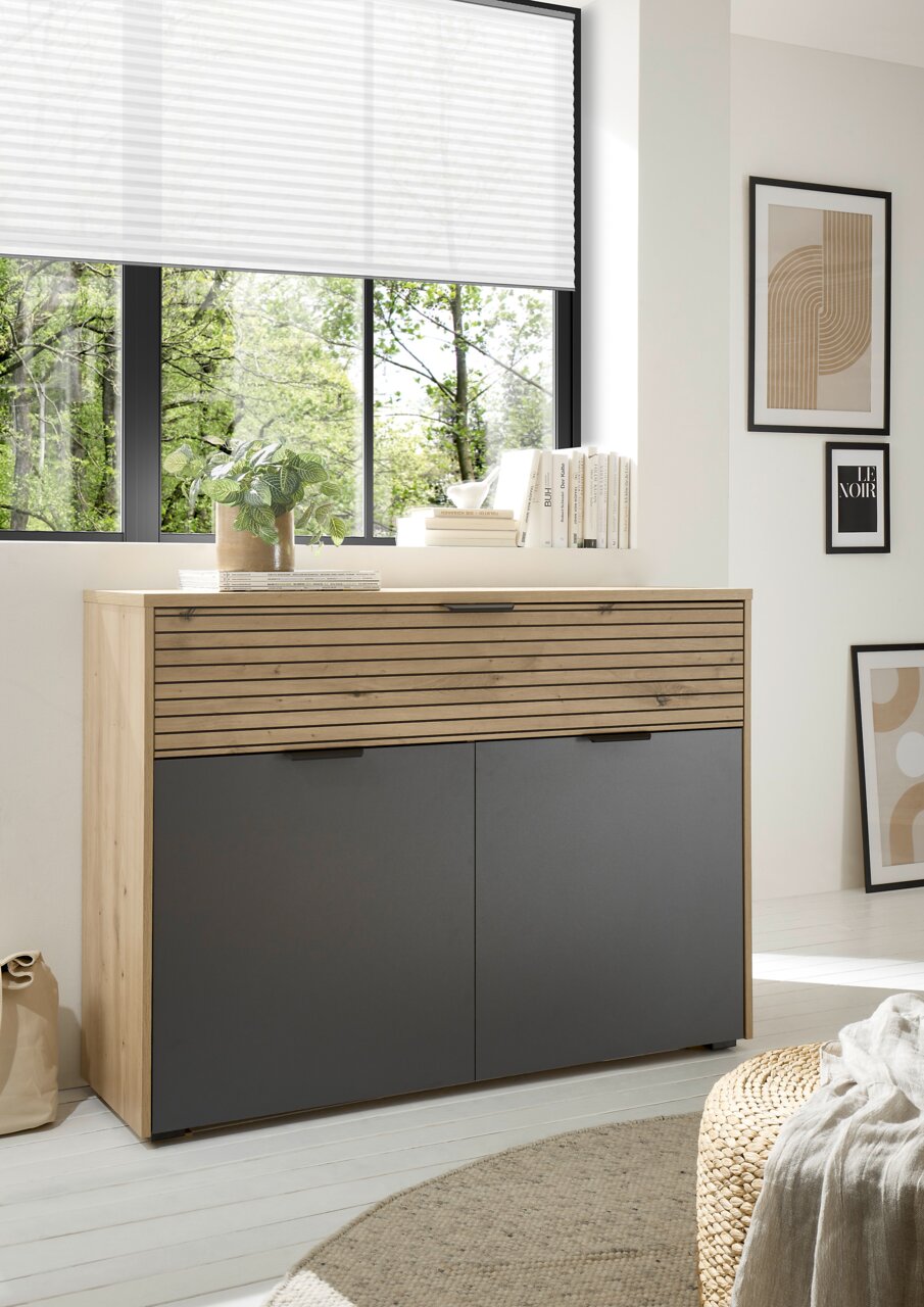Ti´me Kommode STANFORD Moderne Kommode aus Holz mit grauen Türen, fotografiert aus einer leicht seitlichen Perspektive in einem hellen Raum mit großen Fenstern.