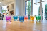 Sechs bunte Trinkgläser in verschiedenen Farben (Pink, Blau, Grün, Orange, Gelb) stehen auf einem Holztisch in einer modernen Küche. Die Perspektive ist frontal auf die Gläser gerichtet.
