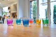Sechs bunte Trinkgläser in verschiedenen Farben (Pink, Blau, Grün, Orange, Gelb) stehen auf einem Holztisch in einer modernen Küche. Die Perspektive ist frontal auf die Gläser gerichtet.