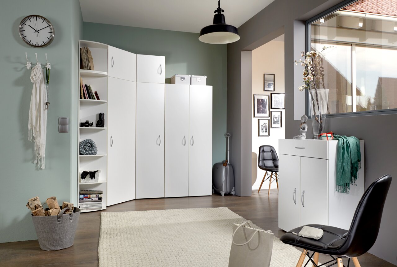 Weiße Mehrzweckschränke und Eckschrank in einem modernen Raum mit grüner Wandfarbe, dekoriert mit einem Teppich und einem Korb mit Holzscheiten. Perspektive von vorne links.