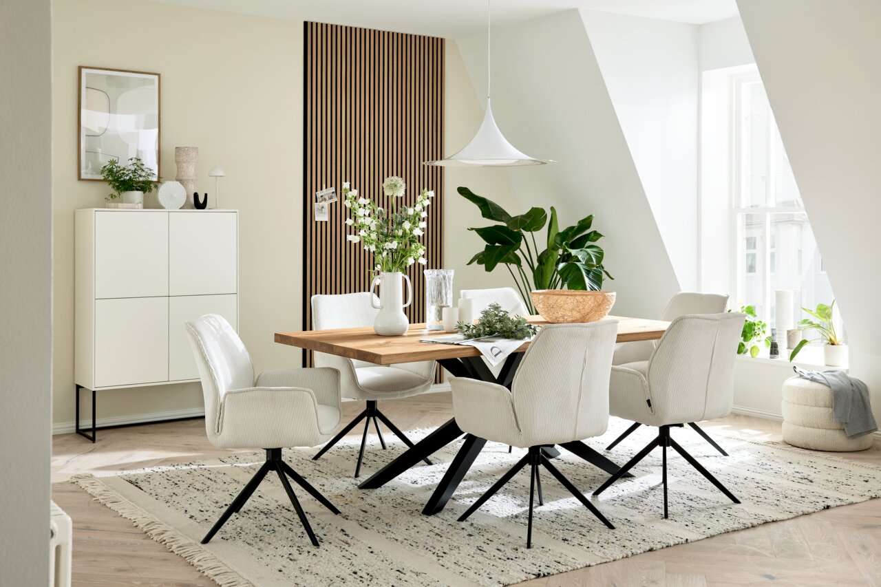 Musterring Fauteuil MR TIAMA Eetkamer met een moderne houten eettafel en witte stoelen met armleuningen, omringd door planten en decoratieve elementen, zijaanzicht.