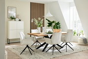 Eetkamer met een moderne houten eettafel en witte stoelen met armleuningen, omringd door planten en decoratieve elementen, zijaanzicht.