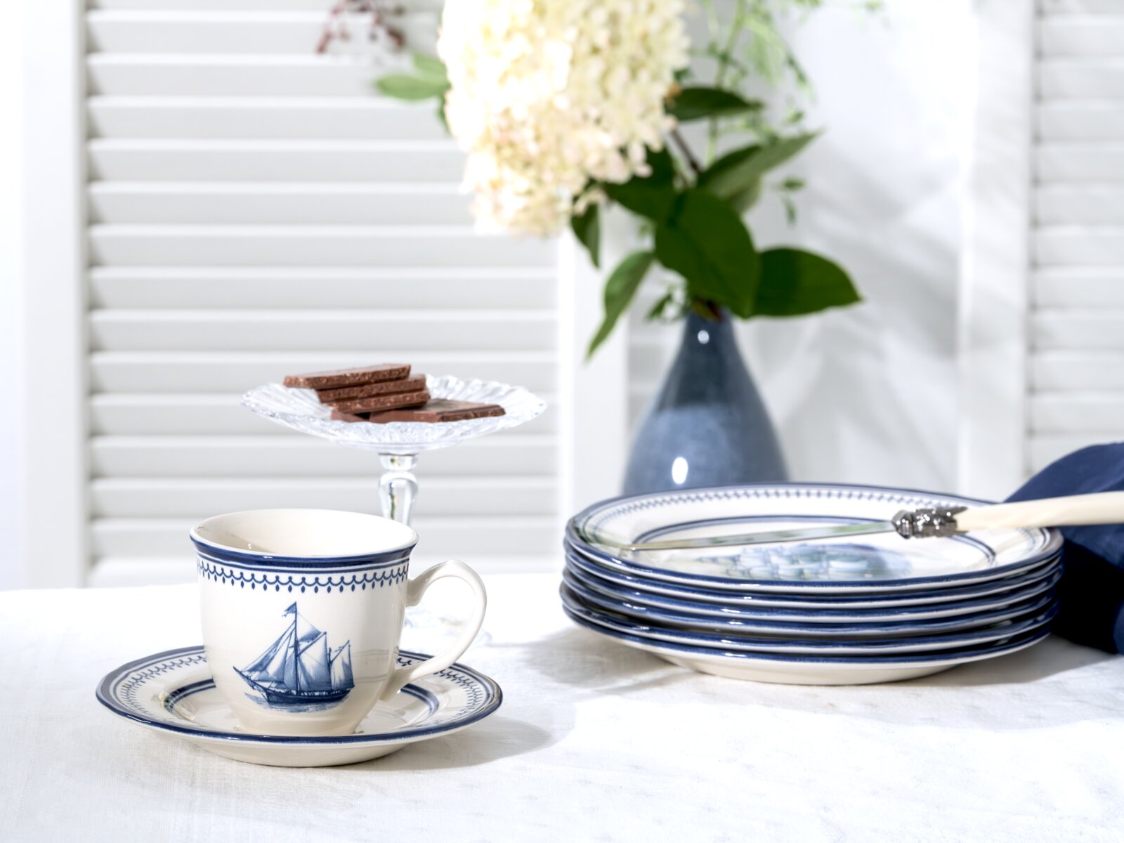 Maritimes Keramik-Geschirr in Weiß-Blau mit Segelschiff-Motiv: Kaffeetasse mit Untertasse und gestapelten Tellern auf weißem Tisch, fotografiert in leicht seitlicher Perspektive auf Tischhöhe; im Hintergrund Glas-Etagere mit Schokolade und blaue Vase mit 