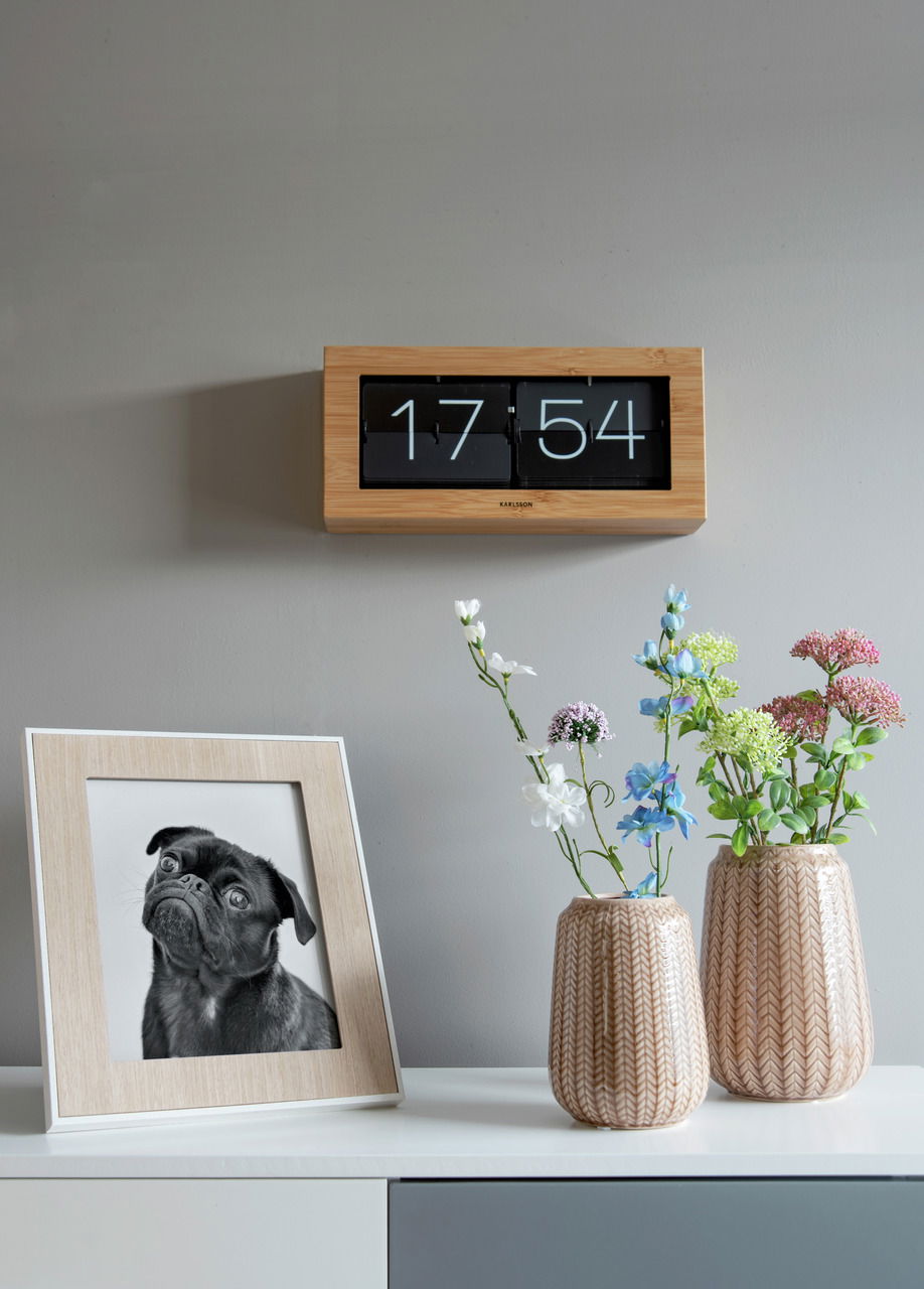 Wanduhr mit Bambusrahmen und Flip-Anzeige, frontal an der Wand montiert, neben einem Tisch mit Blumenvasen und einem gerahmten Foto.