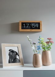 Wanduhr mit Bambusrahmen und Flip-Anzeige, frontal an der Wand montiert, neben einem Tisch mit Blumenvasen und einem gerahmten Foto.