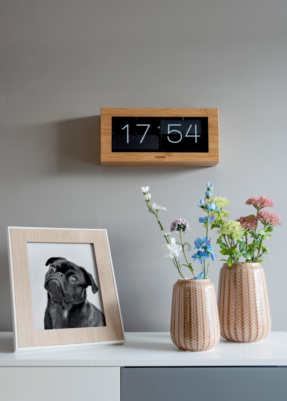 Wandklok met bamboeframe en omklapbaar display, aan de voorkant aan de muur bevestigd, naast een tafel met bloemenvazen en een ingelijste foto.
