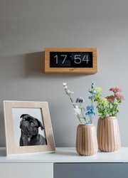Wandklok met bamboeframe en omklapbaar display, aan de voorkant aan de muur bevestigd, naast een tafel met bloemenvazen en een ingelijste foto.