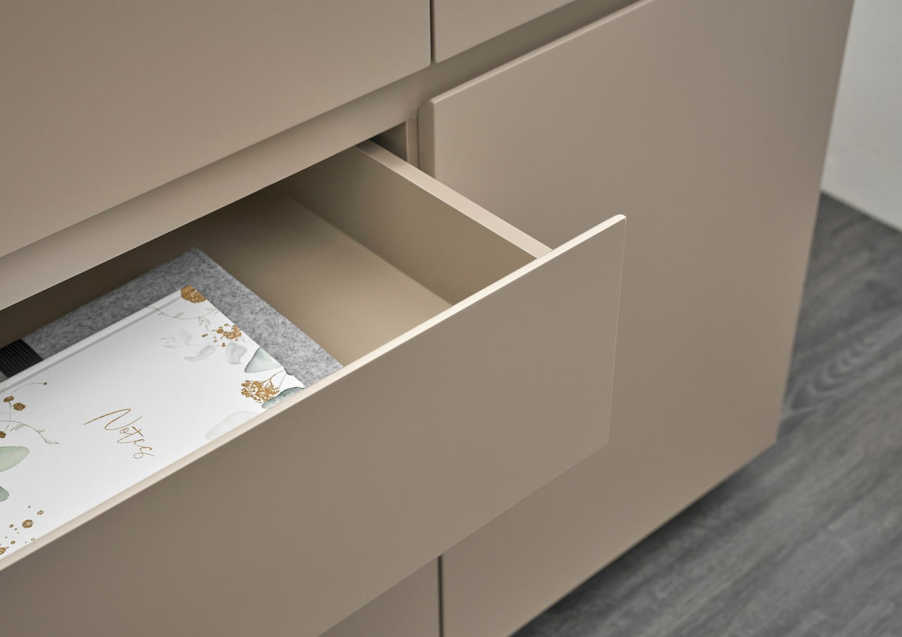 Nahaufnahme einer geöffneten Schublade eines modernen Sideboards in Beige, mit einem Notizbuch und einer Filzunterlage. Perspektive von oben.