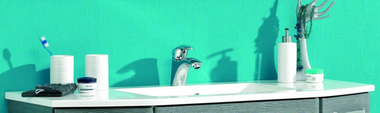 XORA Wastafel van mineraalmarmer ATHEN Moderne wastafel met verchroomde kraan op een wit blad, handdoek en verzorgingsproducten tegen een turquoise muur, vooraanzicht op ooghoogte