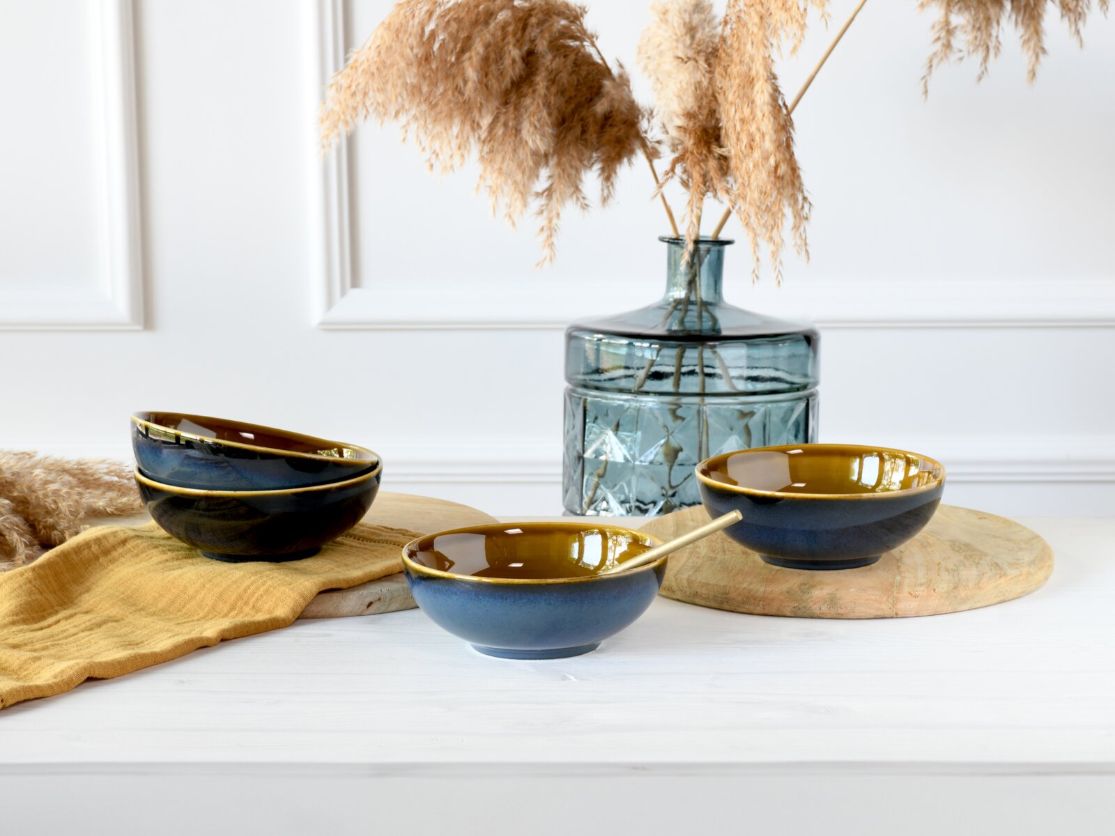 Drei blau-gold glasierte Keramikschalen auf Holzplatten neben einer Glasvase mit Pampasgras, fotografiert in frontaler Perspektive auf Tischhöhe.