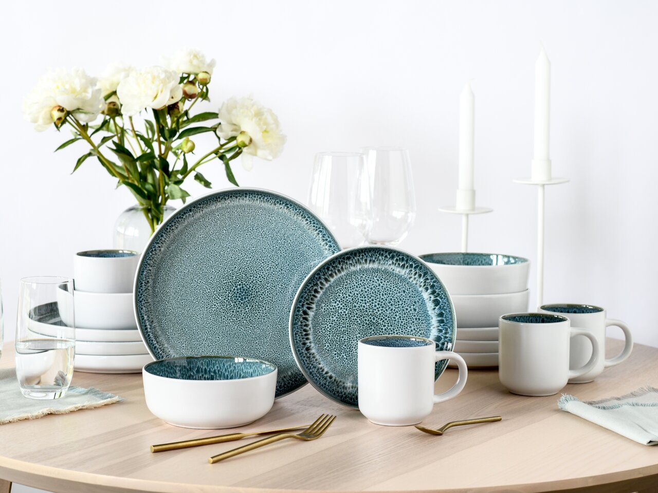 Creatable Kombiservice 16-tlg. NORDIC FJORD Kombiservice 16-tlg. Eisblau auf einem Tisch, bestehend aus Tellern, Schalen und Tassen mit eisblauer Glasur, umgeben von Blumen und Kerzen, aus der Perspektive von vorne.