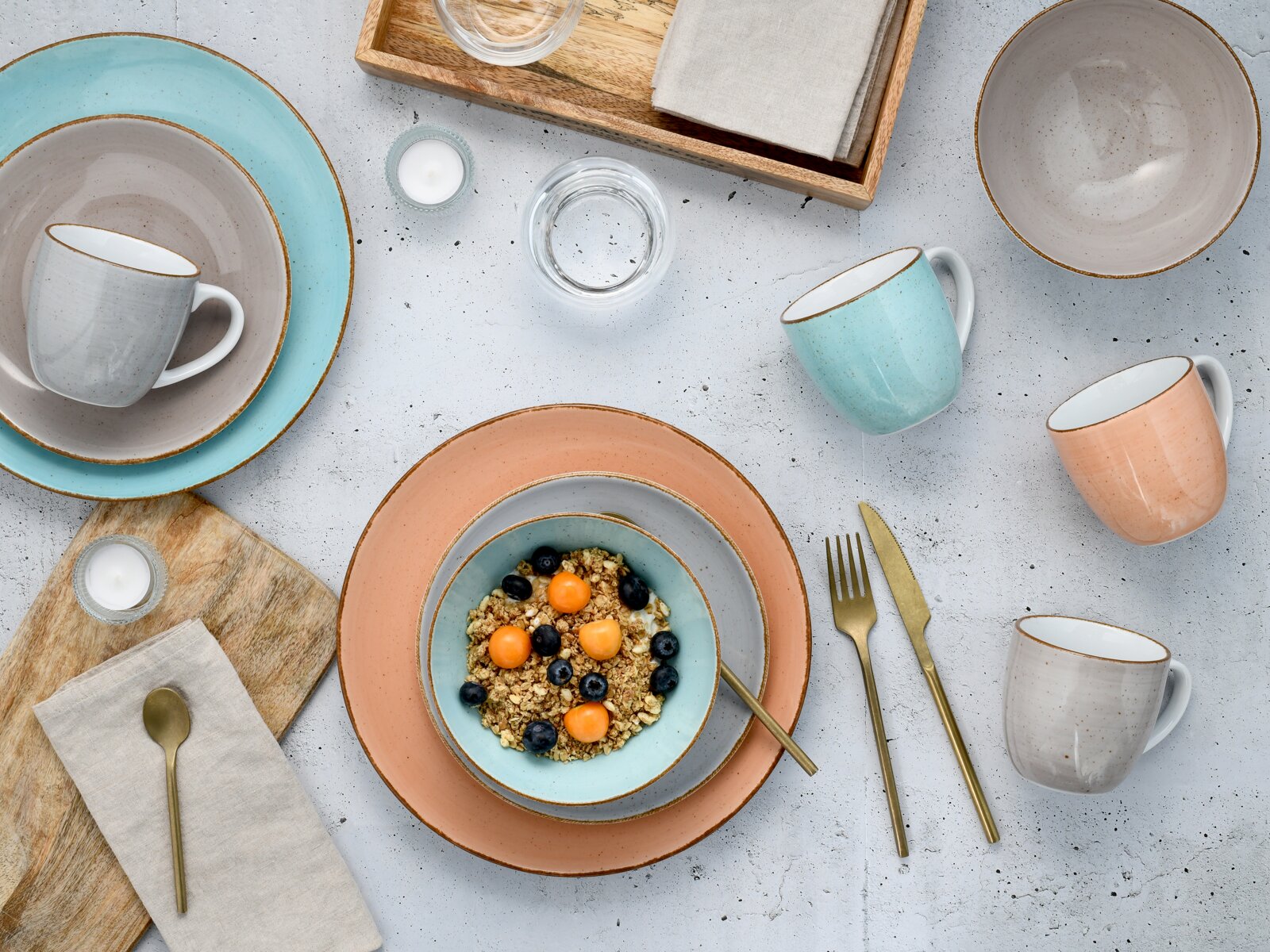 Pastellfarbenes Keramik-Geschirrset mit Tellern, Schalen und Tassen sowie goldfarbenem Besteck auf hellem Tisch, Draufsicht von oben (Flatlay).