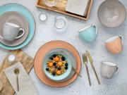 Pastellfarbenes Keramik-Geschirrset mit Tellern, Schalen und Tassen sowie goldfarbenem Besteck auf hellem Tisch, Draufsicht von oben (Flatlay).