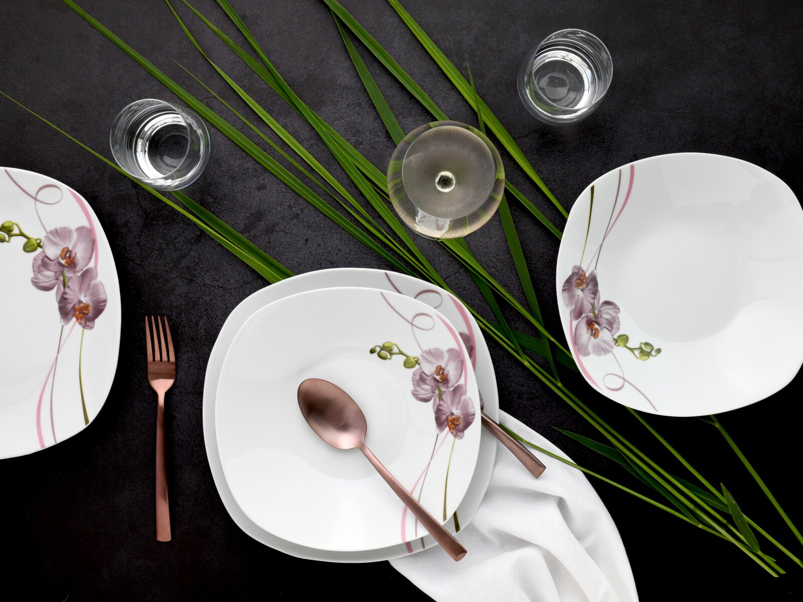 Draufsicht auf ein weißes, quadratisch abgerundetes Tafelservice mit Orchideen-Dekor und rosa Linien, arrangiert mit Roségold-Besteck, Wassergläsern, weißer Serviette und grünen Blättern auf dunklem Tisch.