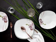 Draufsicht auf ein weißes, quadratisch abgerundetes Tafelservice mit Orchideen-Dekor und rosa Linien, arrangiert mit Roségold-Besteck, Wassergläsern, weißer Serviette und grünen Blättern auf dunklem Tisch.