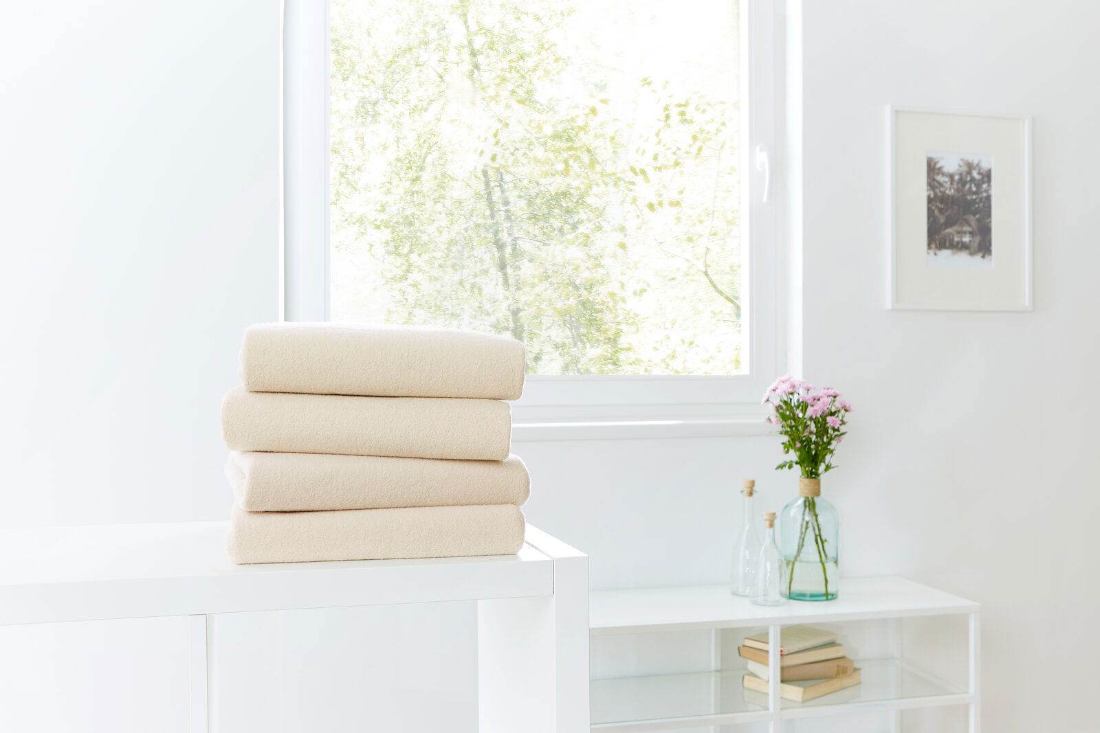 Novel Spannbetttuch TEDDY-FLAUSCH 100x200cm Beige Spannbetttücher gestapelt auf einem weißen Tisch, im Hintergrund ein Fenster mit Blick ins Grüne, seitliche Perspektive.